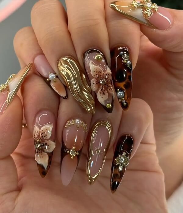 Close-up of long stiletto nails with 3D embellishments, gold accents, floral details, and tattoo-inspired nail art on nude and brown glossy bases.