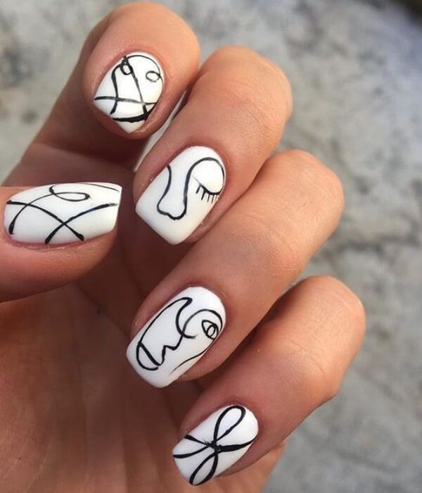 Close-up of hand with white nails featuring abstract black line art face tattoos, minimalist continuous-line designs, and modern artistic nail tattoo style.
