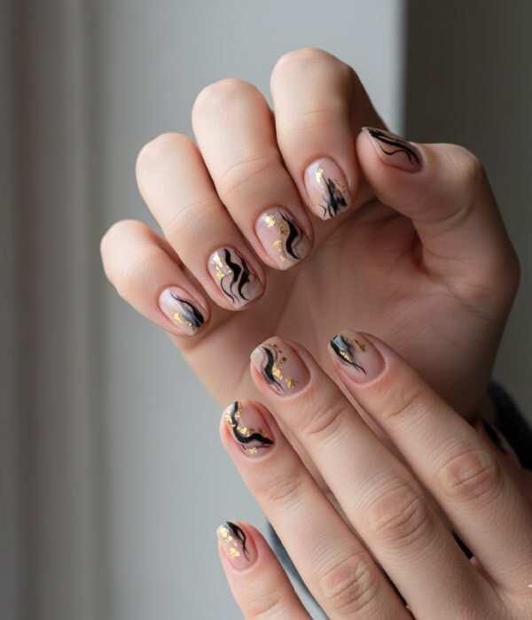 Close-up of nude nails with abstract tattoo designs featuring black flowing lines, watercolor-style swirls, and subtle gold foil accents.