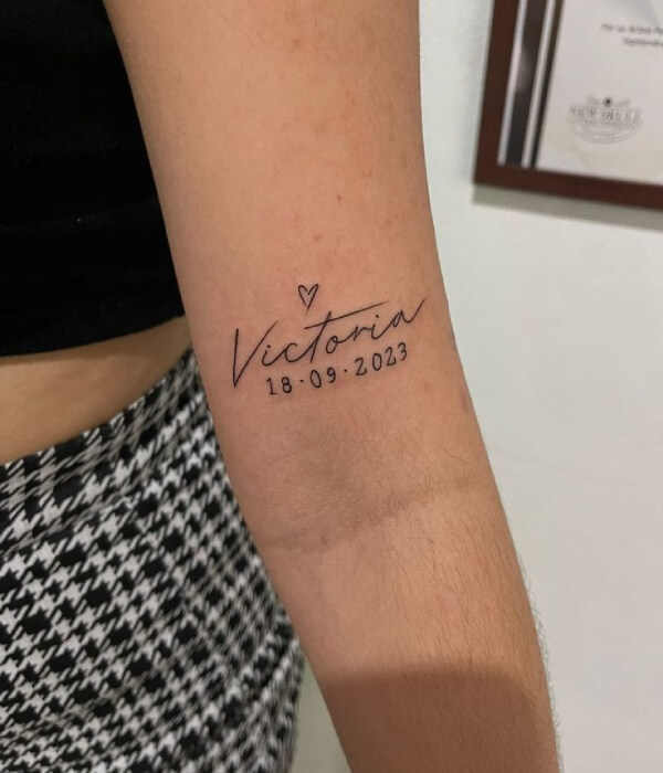 Minimalist name and birthdate tattoo on the forearm featuring the name “Victoria” in fine cursive lettering with a small heart and the date “18-09-2023” in black ink.