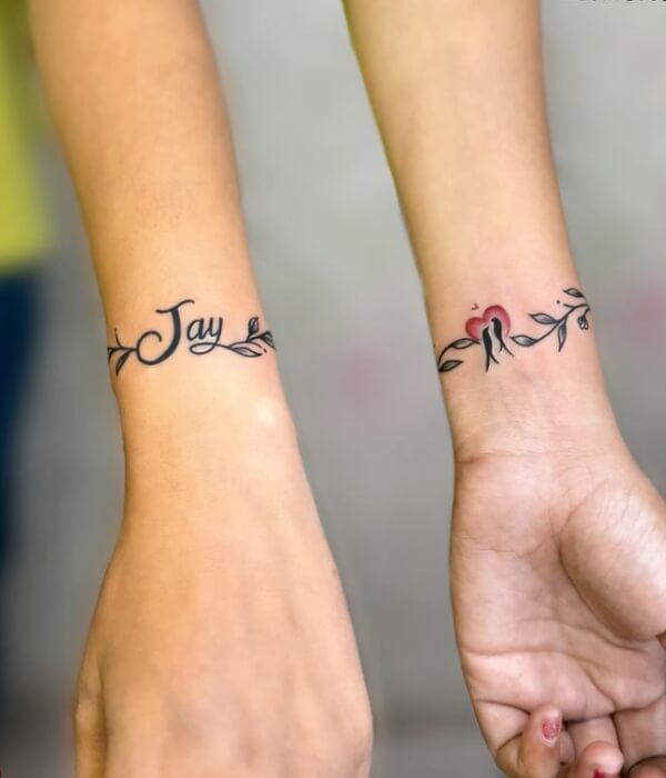 Matching bracelet-style name tattoos on both wrists, featuring the name “Jay” in cursive with delicate leaf accents and a small red heart, inked in black and red.