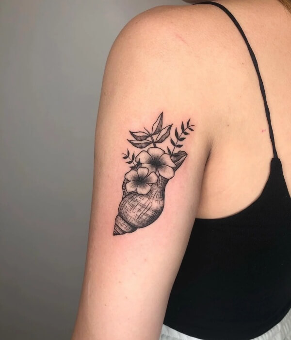 Black ink conch shell tattoo with delicate flowers and leaves on a woman’s upper arm, featuring an elegant nature-inspired shell tattoo design.