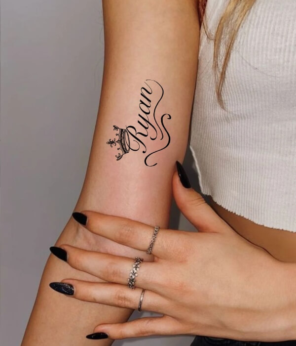 Elegant name tattoo on a woman’s forearm featuring the name “Ryan” in flowing cursive script, accented with a small crown above the lettering in fine black ink.