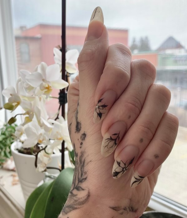 Close-up of natural nude nails featuring delicate floral nail tattoos in fine black line art, with botanical designs extending across the fingertips and a soft indoor background.