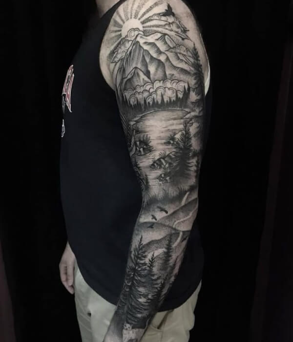 A black-and-grey full-sleeve tattoo of a mountain landscape with a sun, clouds, forest, a lake, and flying birds on a person's arm.