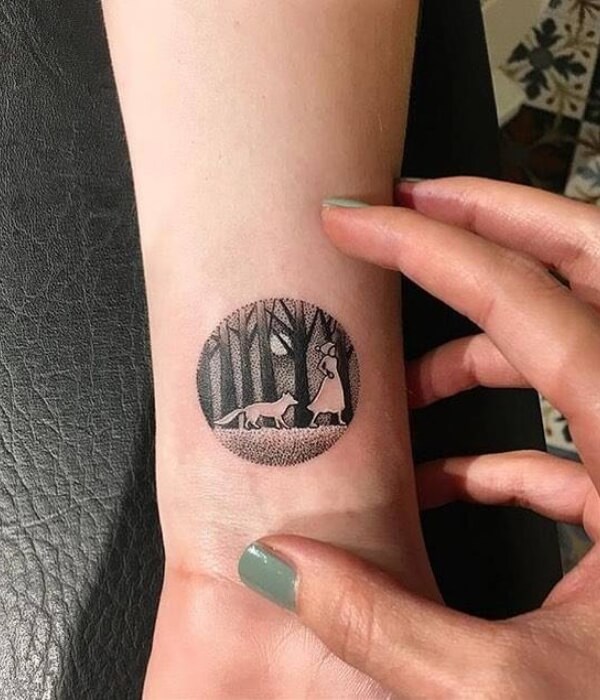 A circular blackwork wrist tattoo featuring a fox and a woman walking through a forest under a full moon.