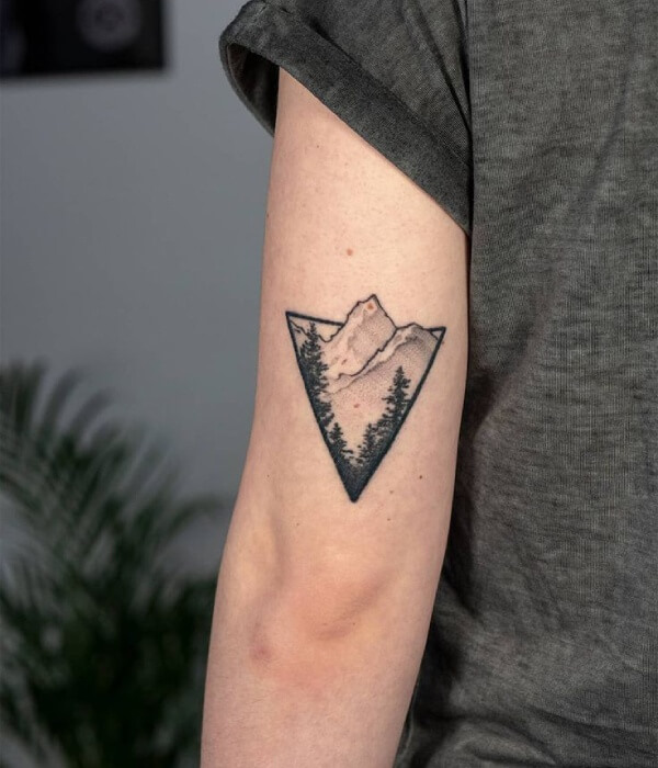 A black-and-grey dotwork tattoo on an upper arm featuring a detailed mountain range above a dense pine forest with three deer in the foreground.