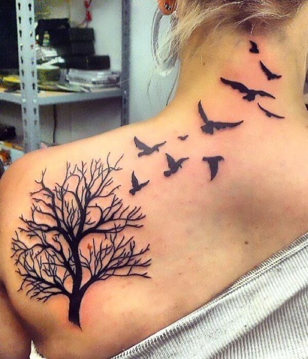 Forest and birds tattoo on the upper back and shoulder, featuring a black silhouette tree with bare branches and flying birds in minimalist black ink on real human skin.