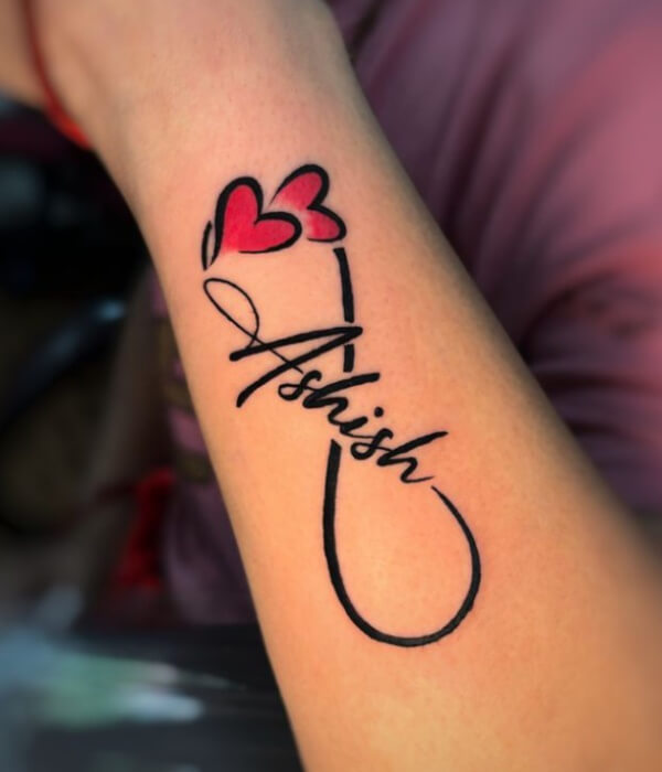 Infinity-style name tattoo on the forearm, featuring the name “Ashish” in bold cursive with an infinity loop and two red heart accents in black and red ink.
