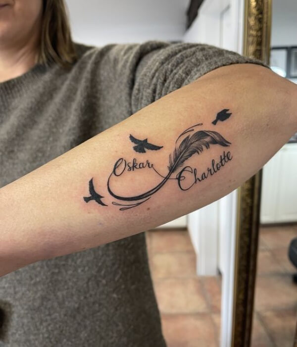 Matching couple name tattoo on the forearm featuring the names “Oskar” and “Charlotte” in elegant cursive, connected by a flowing feather design with small flying birds in black ink.