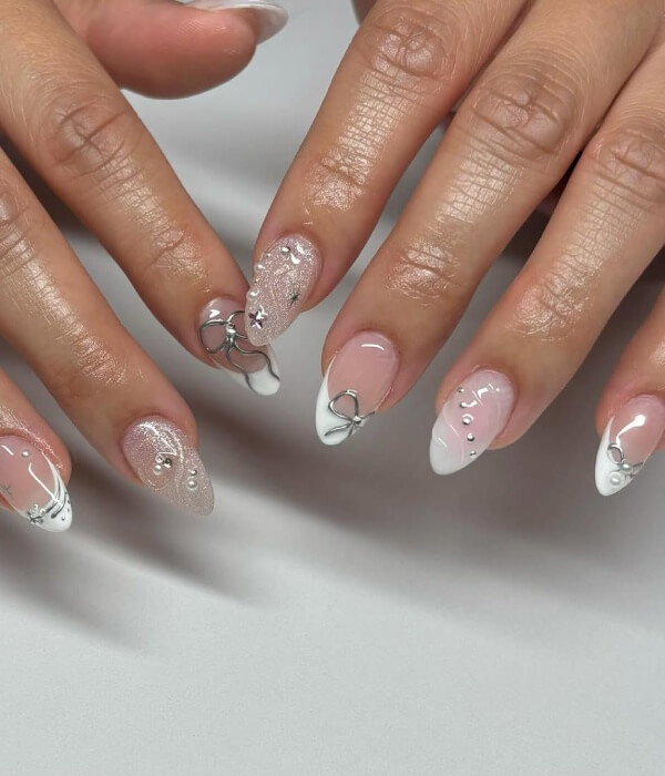Close-up of almond-shaped nude and white nails with metallic accent tattoo designs, silver wire details, rhinestones, and a soft glossy finish.