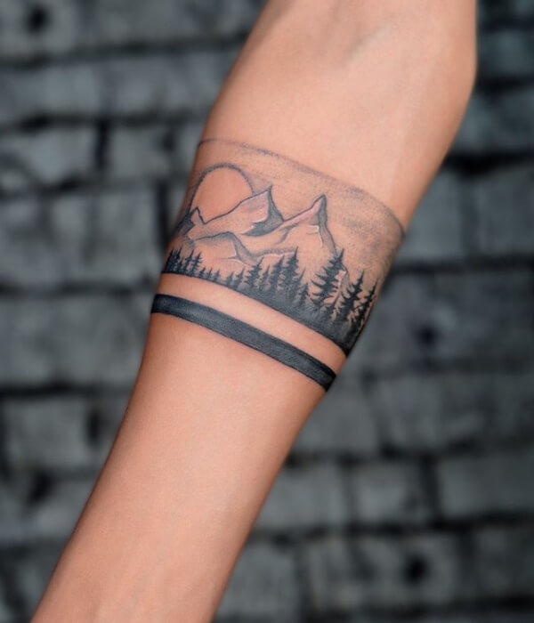 Minimalist forest band tattoo on an arm, featuring fine-line mountains, pine trees, and a solid black armband in subtle black and grey ink on real human skin.