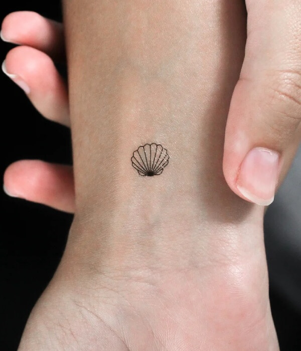 Tiny minimalist scallop shell tattoo on the wrist featuring a delicate black line seashell design.