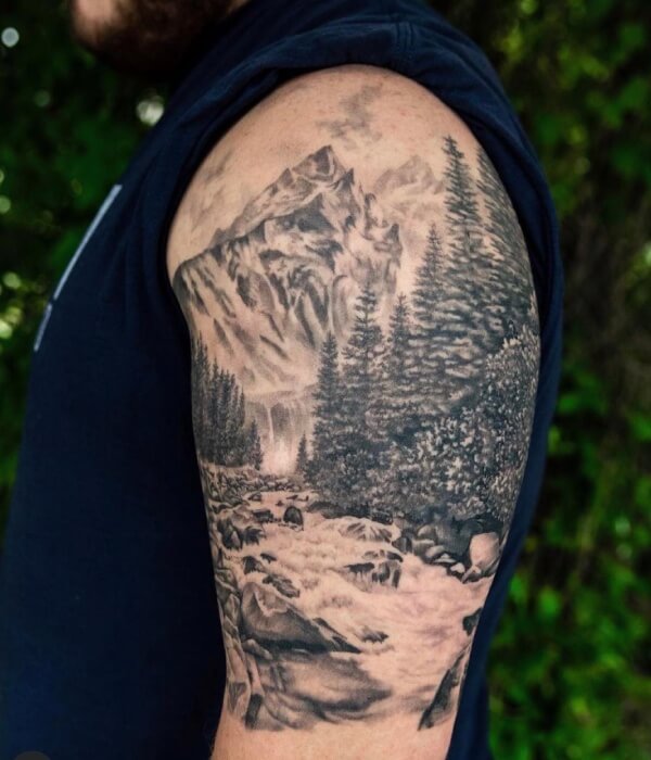 Realistic mountain forest landscape tattoo on an upper arm, featuring detailed pine trees, rocky mountains, and a flowing river in black and grey realism style on natural human skin.