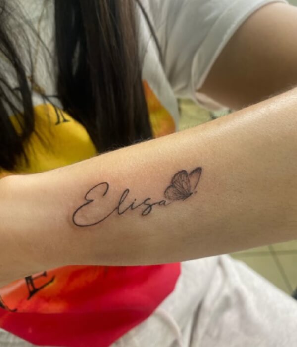 Calligraphy-style name tattoo on the forearm featuring the name “Elisa” in elegant cursive lettering, accented with a small butterfly in fine black ink.
