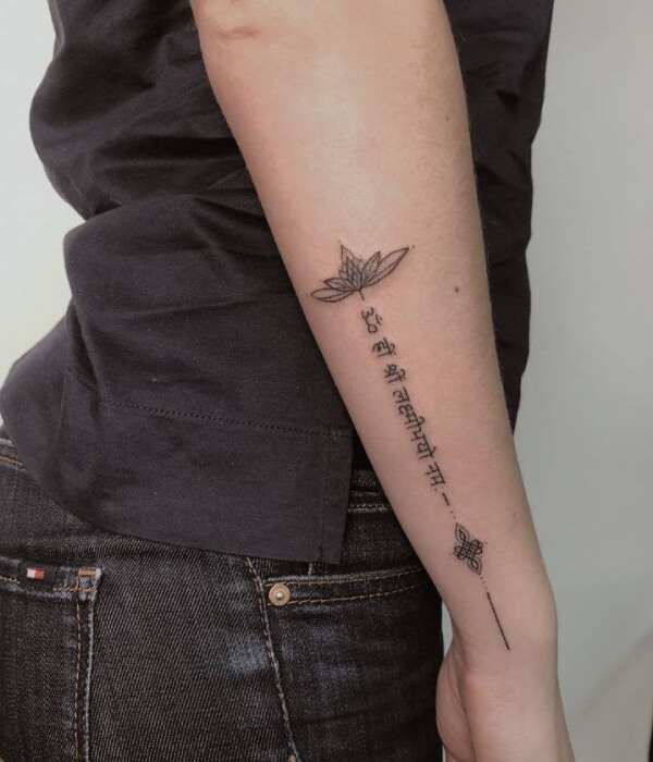 Vertical Sanskrit script name tattoo on the inner forearm, featuring Devanagari lettering with a lotus flower at the top and a minimalist symbol at the bottom, inked in fine black lines.
