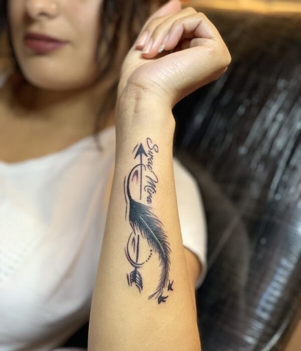 Name tattoo on a woman’s forearm featuring elegant cursive lettering paired with a feather and arrow design, accented with fine line details and small birds in black and blue ink.