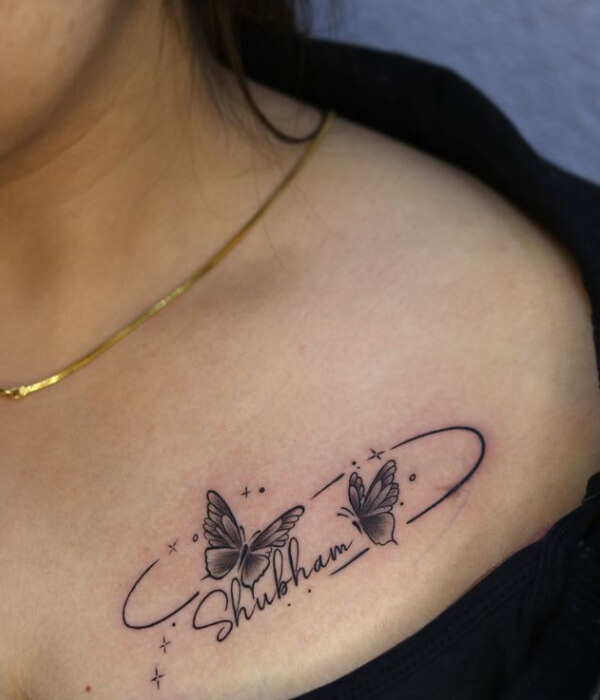 Name tattoo on the collarbone featuring the word “Shubham” in elegant cursive, accented with two detailed butterflies, fine linework, and small star elements in black ink.
