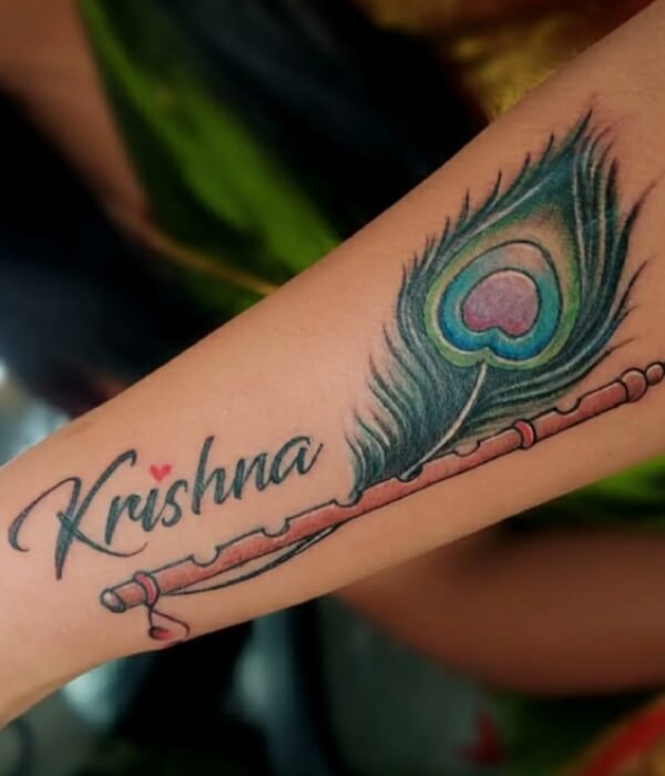 Name tattoo on the forearm featuring the word “Krishna” in elegant script, paired with a detailed peacock feather and flute design in vibrant green, blue, and brown tones.