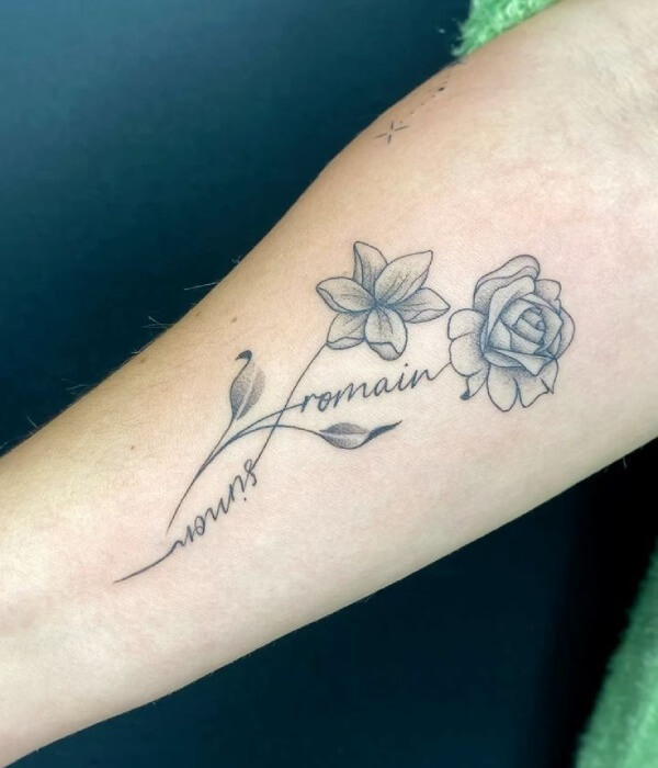 Floral name tattoo on the forearm featuring the name “Romain” in cursive script, accented with delicate line-art flowers and leaves in fine black and grey shading.