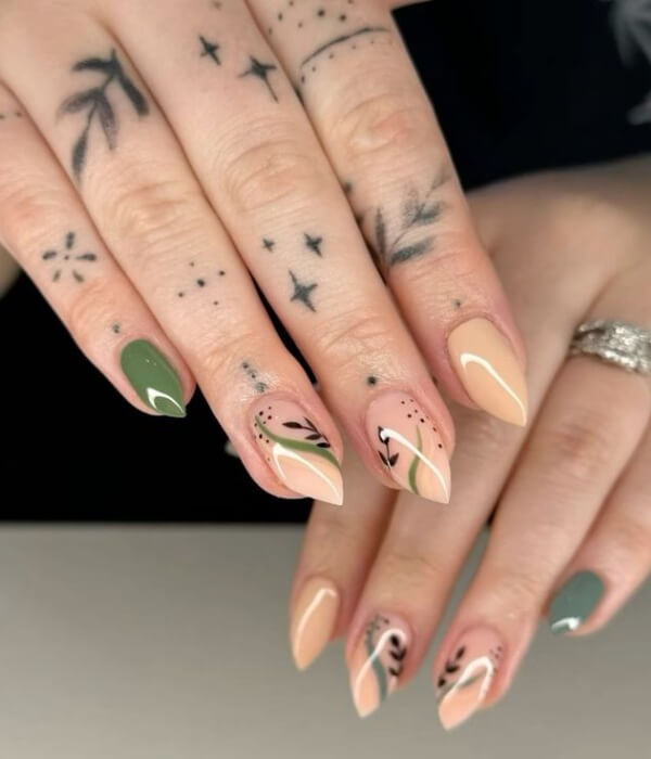 Close-up of almond-shaped nude and sage green nails with nature-inspired tattoo designs, minimalist leaf motifs, fine-line accents, and subtle star details.