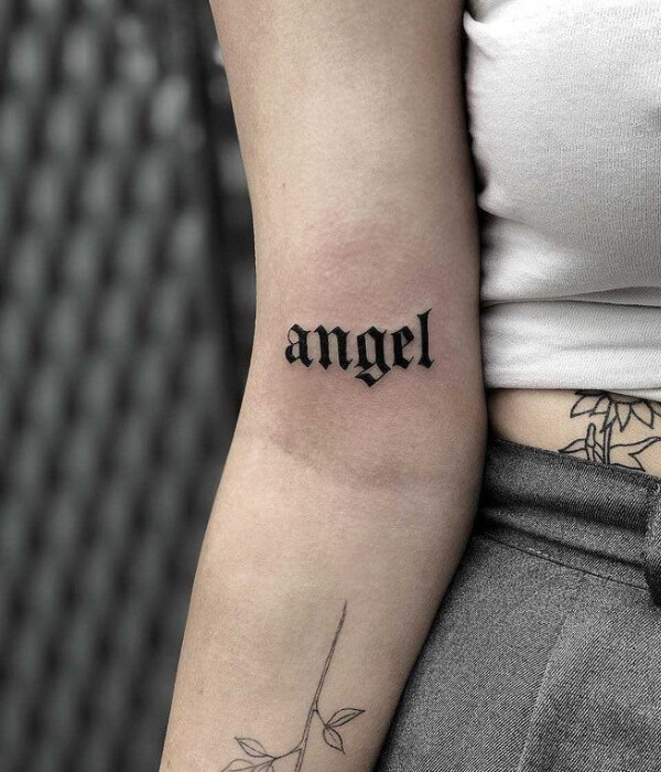 Old English font name tattoo reading “angel” in bold black lettering on the inner arm, featuring a classic gothic style with clean lines and minimal surrounding detail.
