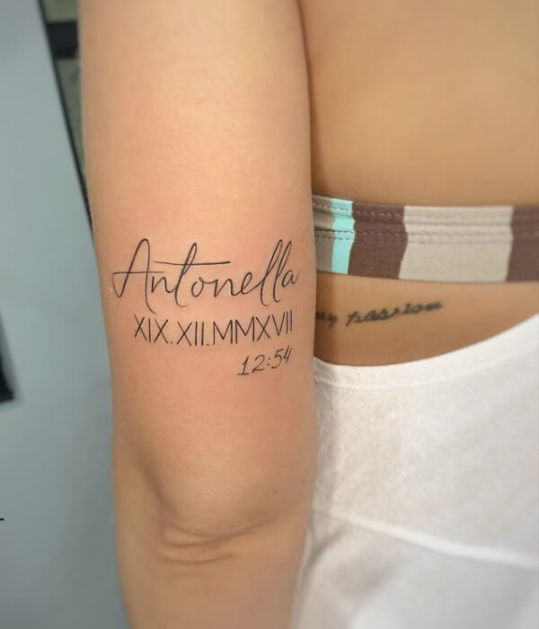 Minimalist name tattoo on the inner arm featuring the name “Antonella” in elegant cursive, paired with Roman numerals and a small time detail, inked in fine black lines.