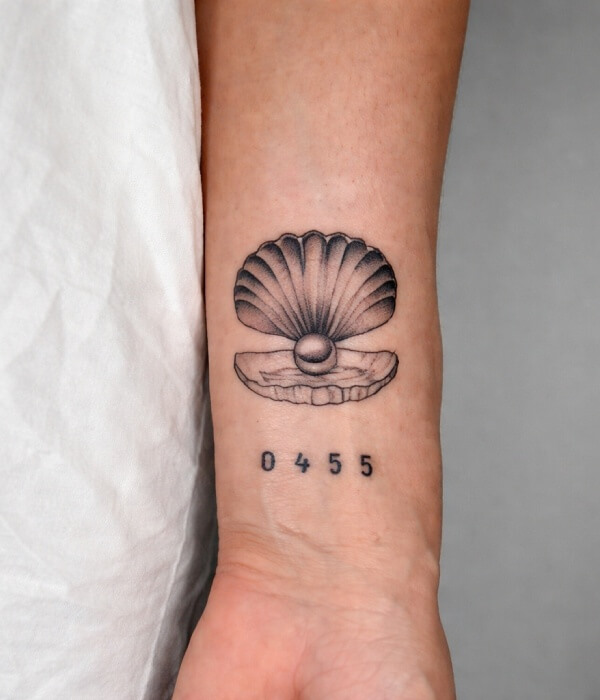 Black and grey scallop shell tattoo with a pearl on the inner forearm, featuring a detailed seashell design with numbers inked below.