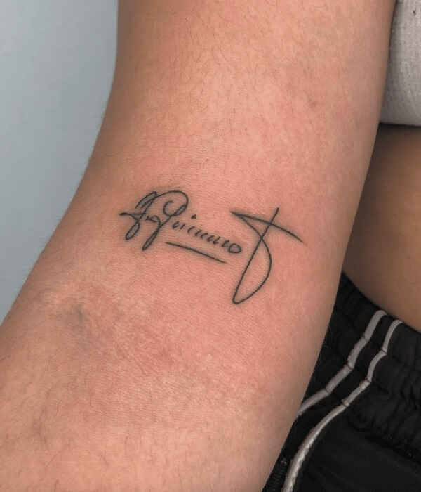 Signature-style name tattoo in black ink on the inner arm, featuring a handwritten cursive name with an underline and flowing, minimalist linework.
