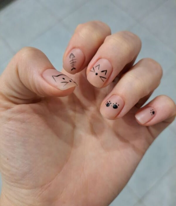 Close-up of nude nails featuring tiny animal motif tattoo designs, including cat faces, fish bone, paw prints, and minimalist black line art.