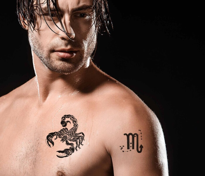 Tiny Zodiac Chest Tattoo 1 - Trending Tattoo Muscular man with a bold black Scorpio zodiac tattoo on the chest and a minimalist Scorpio symbol inked on the shoulder, photographed against a dark background.