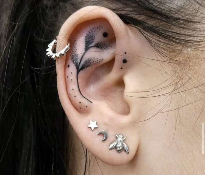 Tiny Zodiac Tatto as ear caffs - Trending Tattoo Close-up of an ear with a delicate black ink leaf tattoo inside the ear cartilage, accented with small dot details and paired with moon, star, and bee earrings.