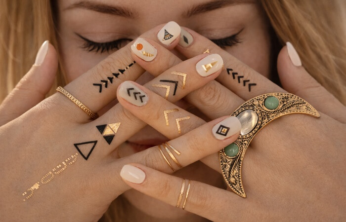 Close-up of hands covering a woman’s face, featuring nude nails with minimalist nail tattoos, black and gold geometric symbols, arrow designs, tiny leaves, and delicate rings in a soft editorial style.