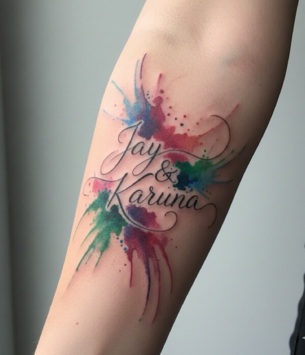 Watercolor name tattoo on the forearm featuring the names “Jay & Karuna” in elegant cursive, surrounded by vibrant splashes of blue, red, green, and purple watercolor ink on skin.