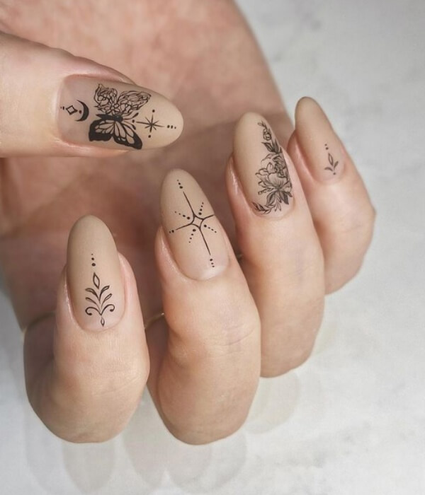 Close-up of nude beige nails featuring delicate black nail tattoos, including a butterfly, floral line art, starburst, and minimalist botanical designs.