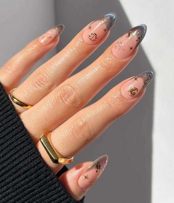 Close-up of almond-shaped nude nails with zodiac sign tattoo designs, gold star details, and subtle glitter tips in a celestial nail art style.