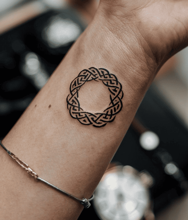 Minimalist black ink circular knot tattoo on the inner wrist, featuring an interwoven Celtic-style design on skin with a blurred background.