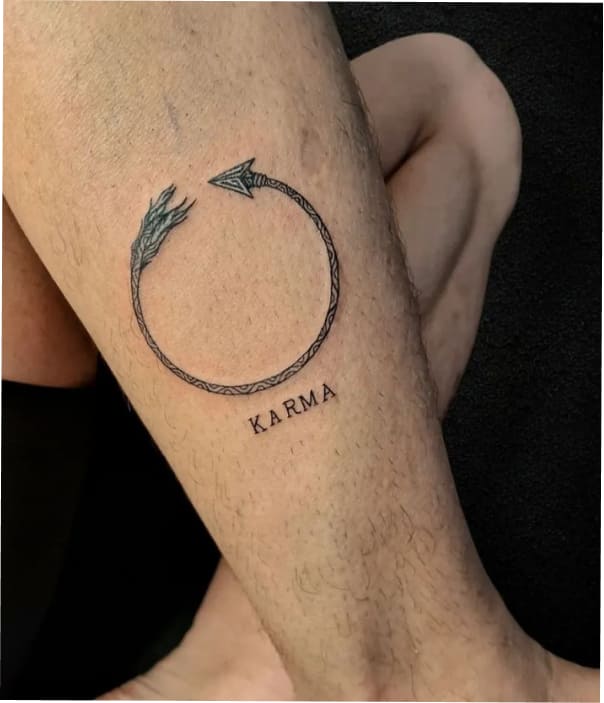 Minimalist forearm tattoo showing a circular arrow with a feathered tail and arrowhead, symbolizing karma, with the word “KARMA” written below in simple lettering.