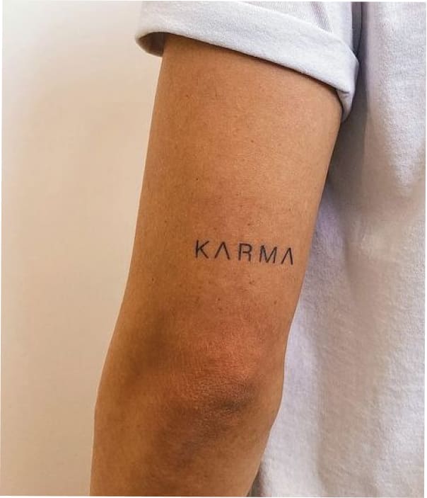 Simple black “KARMA” text tattoo on the inner arm, placed just above the elbow, in clean minimalist lettering.