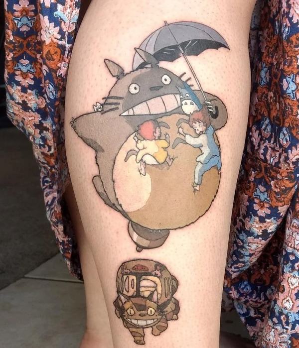 A colorful leg tattoo featuring characters from Studio Ghibli's "My Neighbor Totoro," showing Totoro holding an umbrella with Satsuki and Mei clinging to him, and the Catbus positioned below.