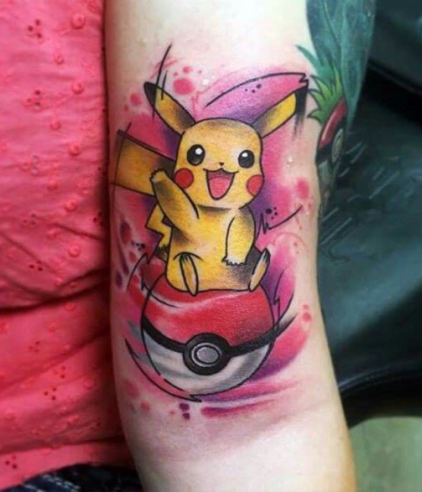 A vibrant, colorful arm tattoo of the Pokémon Pikachu sitting atop a classic red and white Poké Ball. The design features a dynamic "watercolor" style background with splashes of pink and purple ink.