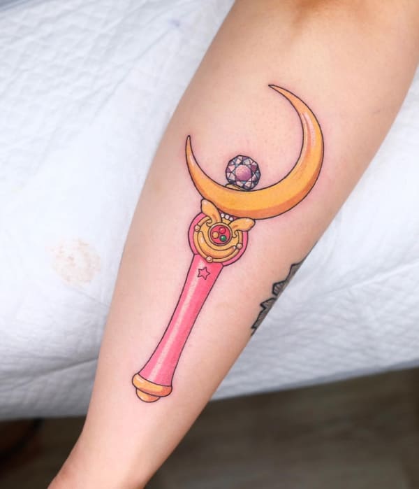 A vibrant, colorful tattoo of Sailor Moon’s Moon Stick on a forearm, featuring a pink handle, a golden crescent moon, and a sparkling purple gem.